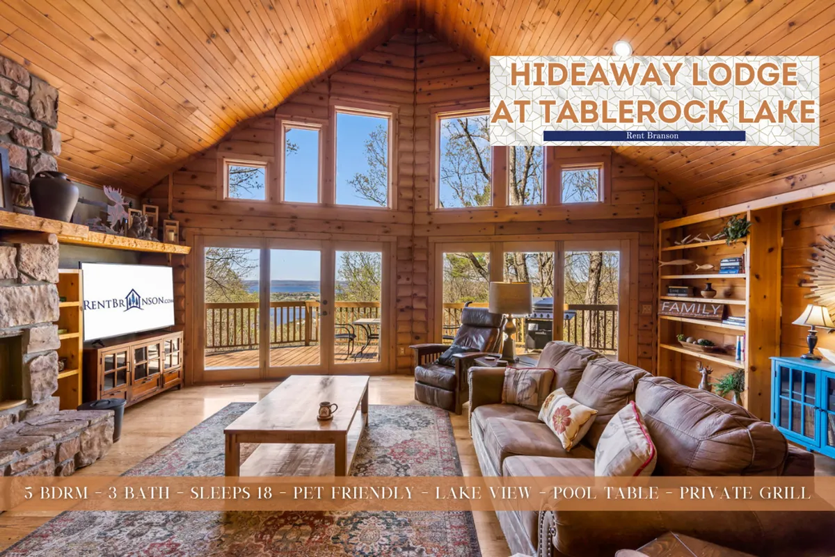 A spacious great room with a striking stone fireplace, large windows that offer a view of the lake, plush, comfortable seating, a wooden coffee table, and a pool table in the background. Natural light streams through the windows, bathing the room in a warm glow and drawing attention to the inviting communal area.