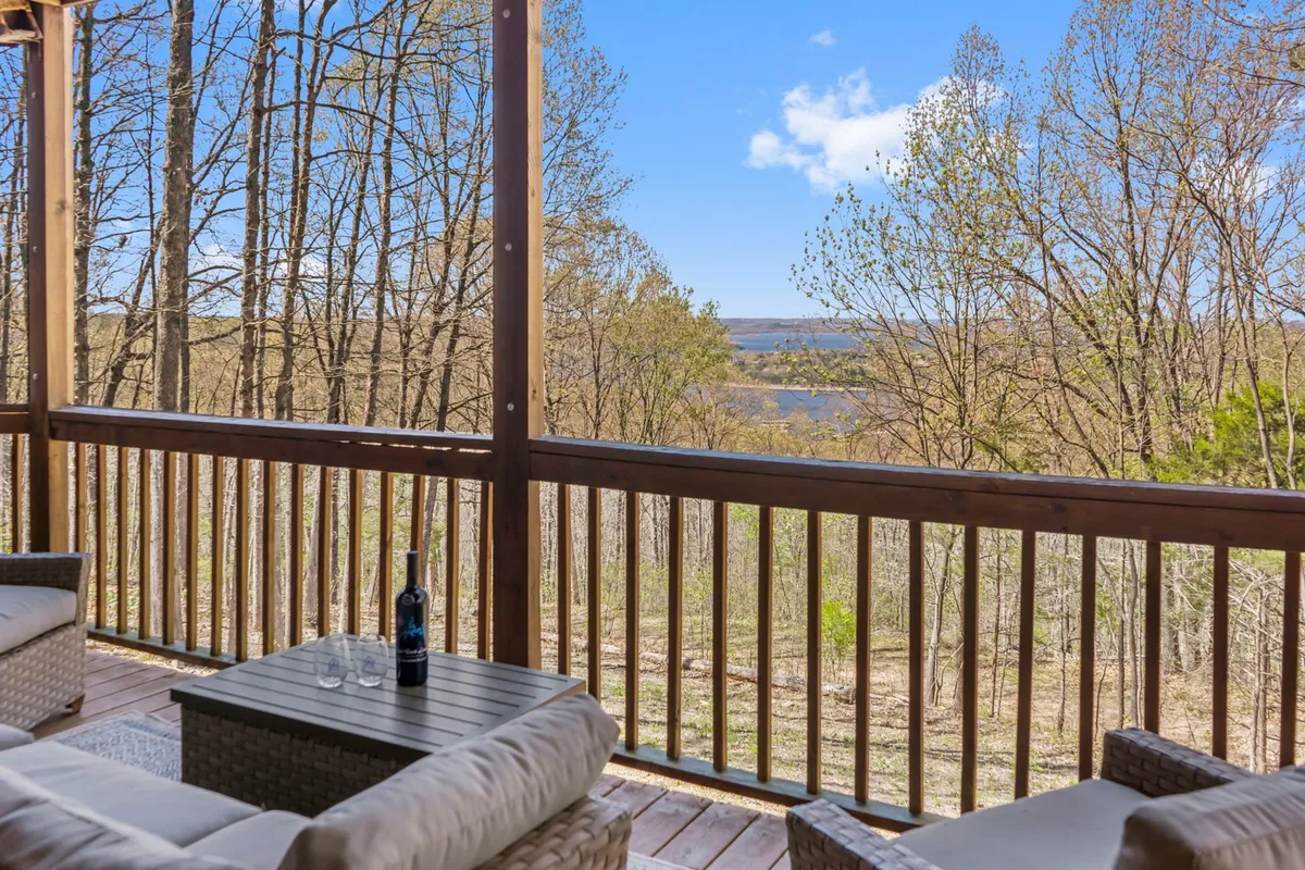 A spacious deck outfitted with comfortable furniture overlooks a stunning lake framed by lush trees. The tranquil setting is perfect for outdoor relaxation, with clear views of the water and surrounding nature. The area is bathed in natural light, inviting guests to enjoy drinks, conversation, or quiet reflection while soaking in the scenery.