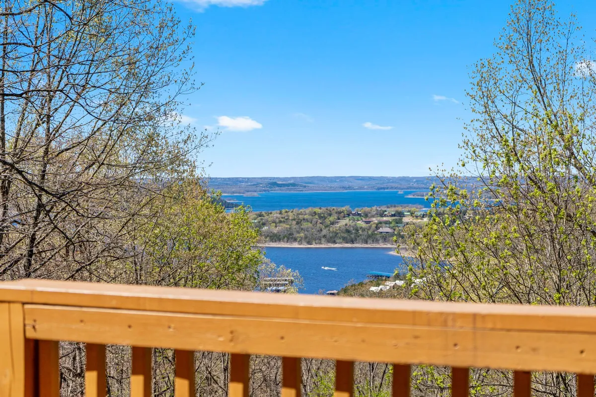 A serene shot from the perspective of someone relaxing on the deck, looking out over an expansive lake view, distant hills, and a tree line. The wooden railing frames the scene, emphasizing the close connection to nature.