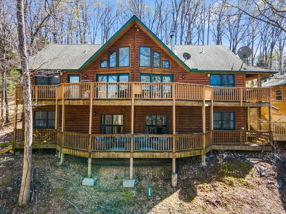 A spacious wooden deck attached to a log cabin, surrounded by tall trees and large windows. The deck is set up for outdoor gatherings and overlooks a serene natural landscape, perfect for family barbecues, morning coffee, or stargazing at night.