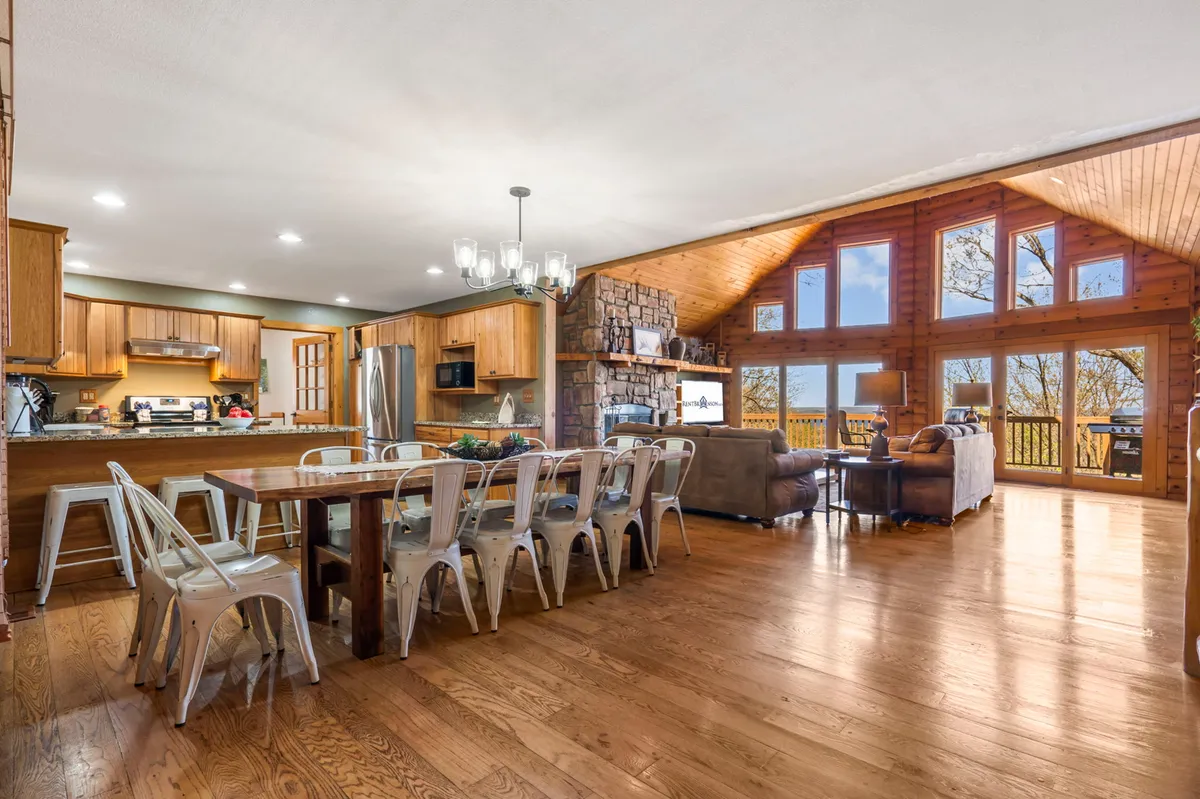 The image features a welcoming dining area with a large wooden table set in front of expansive windows, a prominent stone fireplace, and high ceilings. The arrangement is perfect for family meals or special gatherings, with plenty of natural light and a warm, homey atmosphere.
