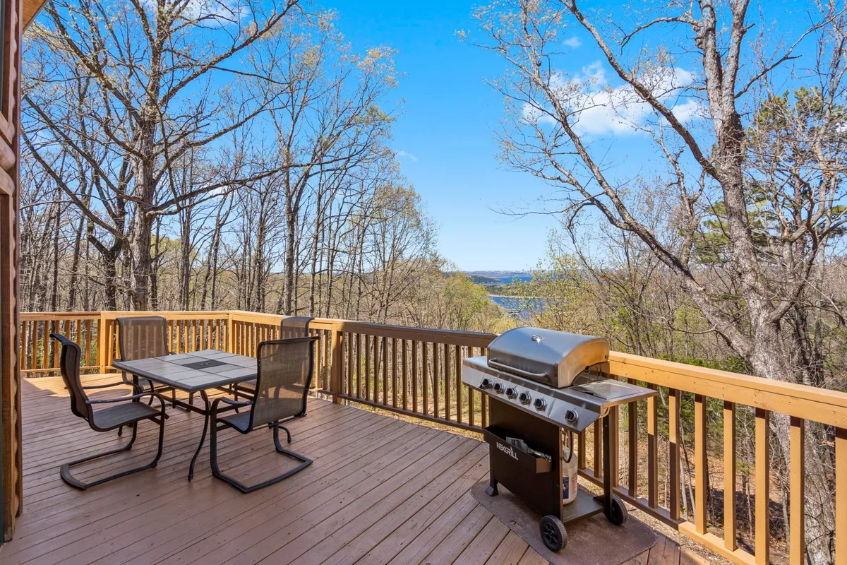 This image features an outdoor dining table set on a deck, complete with a grill and sweeping views of the lake in the background. The deck is bathed in natural light and is surrounded by trees, creating an inviting space for outdoor gatherings or tranquil relaxation.