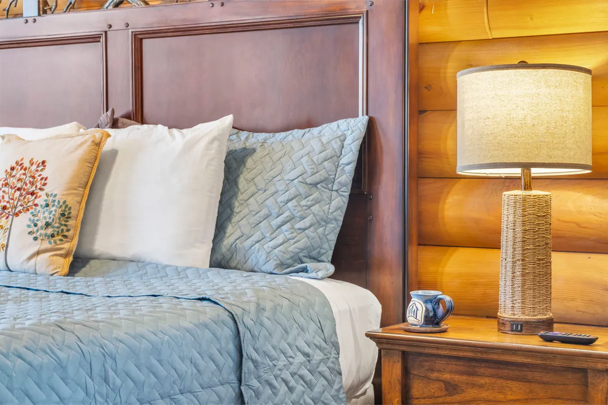 A tranquil bedroom with a bed dressed in a blue quilted coverlet, floral decorative pillow, a wooden nightstand, a woven-texture lamp, and a ceramic mug. Sunlight softly brightens the room, creating a peaceful retreat.