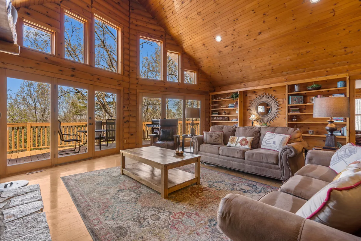 The image showcases a warm and inviting living area inside a rustic log cabin, featuring expansive windows framing serene wooded views, plush seating perfect for relaxing, and charming wooden decor that exudes coziness. A decorative bookshelf hints at quiet moments by the fire, while the spacious layout suggests plenty of room for family gatherings.